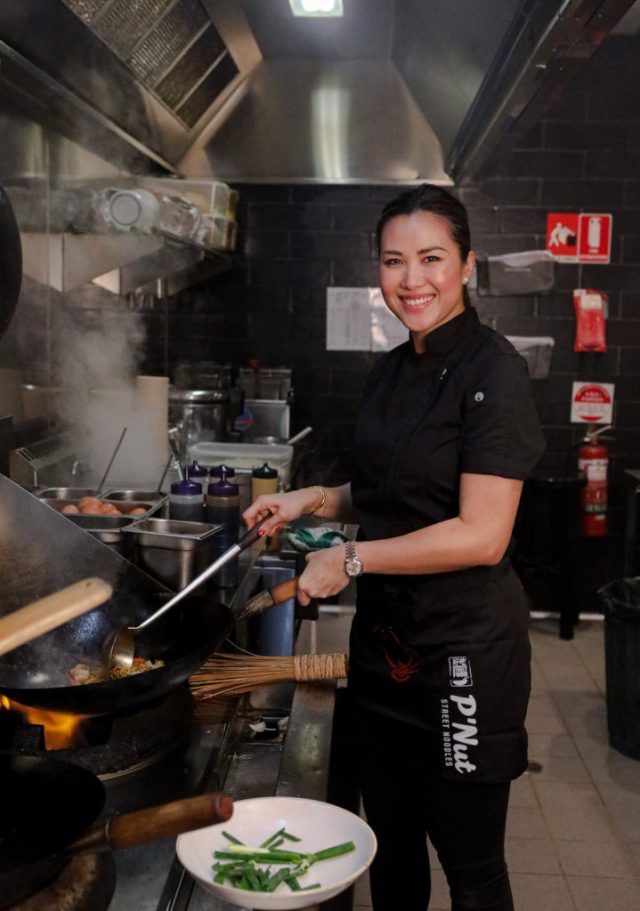 Diana Chan in black chef clothes standing in a commercial kitchen using a wok on an open flame.